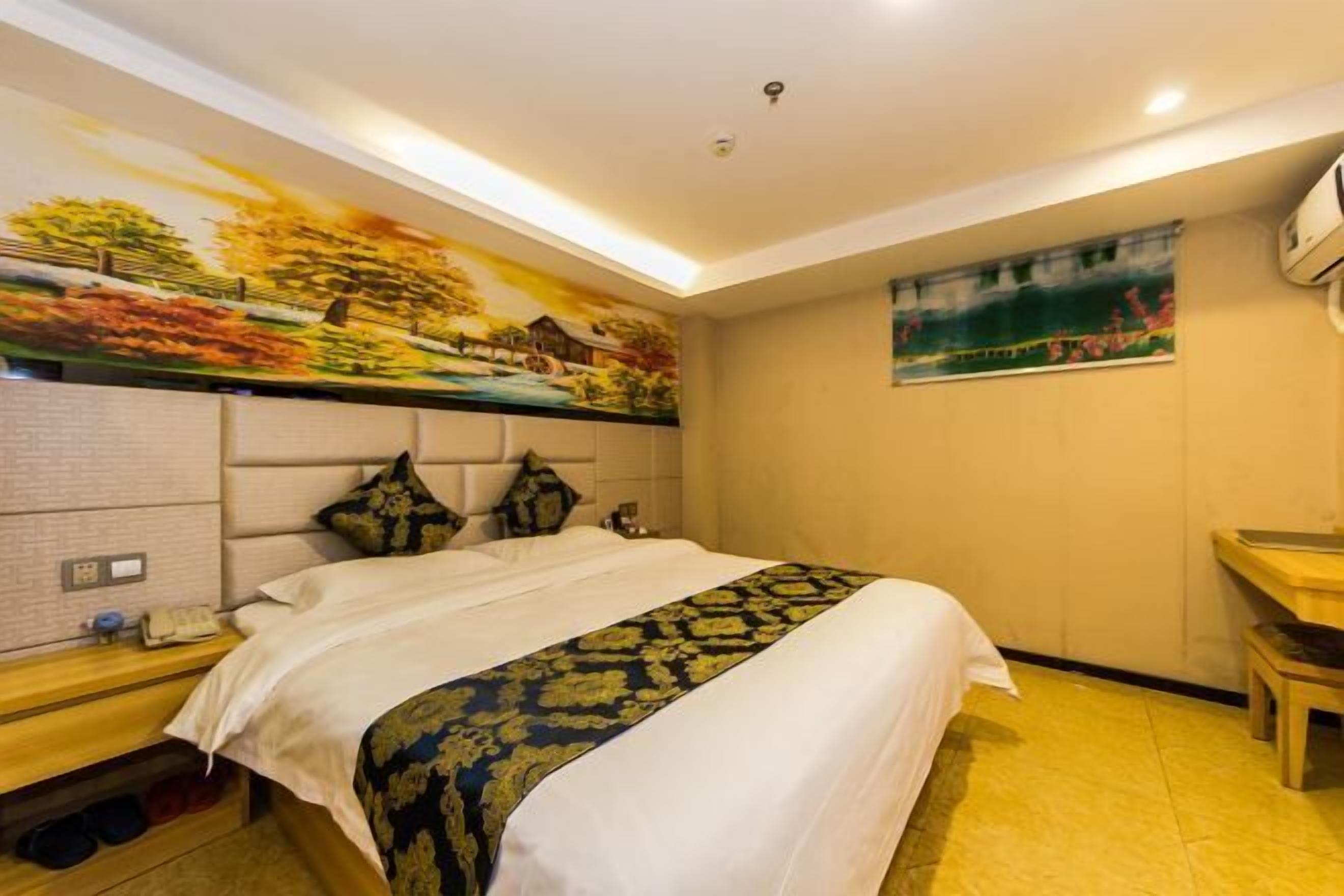 A hotel room with a bed, a headboard with a painting, a bedside table with a phone, and a wall-mounted air conditioner.