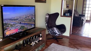 A living room with a television displaying a cityscape, a wooden entertainment unit with bottles, a modern chair, and a large mirror reflecting the room. - Paradise on the shores of Ibiraquera Lagoon (Imbituba)