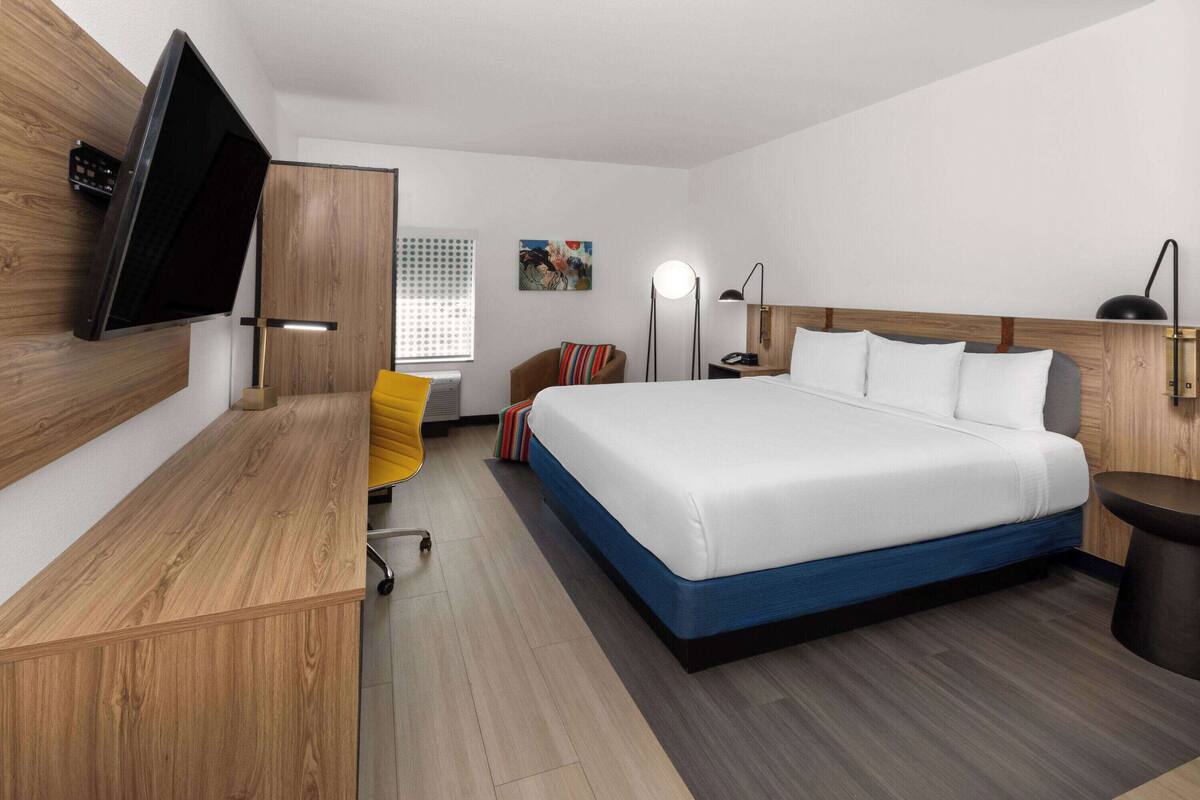 Ein modernes Hotelzimmer mit einem großen Bett, einem Schreibtisch, einem Sessel, einem an der Wand montierten Fernseher und einer Nachttischlampe.