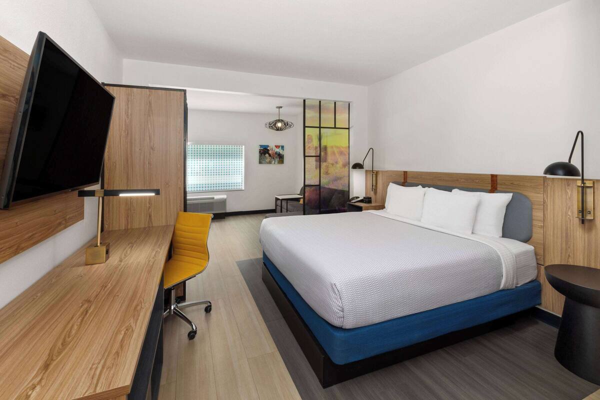 Ein modernes Hotelzimmer mit Bett, Schreibtisch, Stuhl, Fernseher und einem Fenster mit Jalousien.