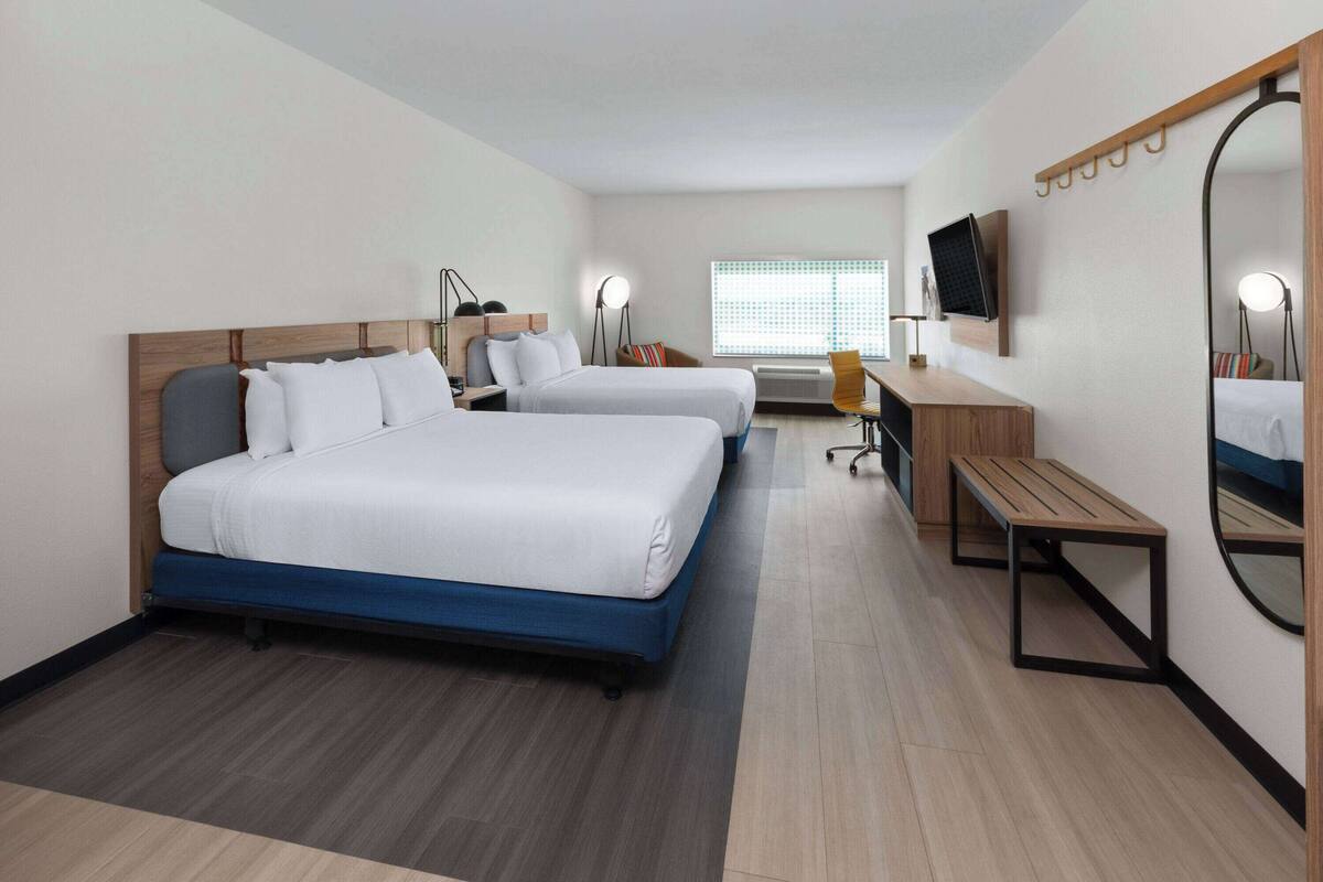 Ein Hotelzimmer mit zwei Betten, einem Schreibtisch, einem Stuhl, einem Fernseher, einem Spiegel und einem Fenster mit Jalousien.