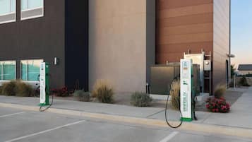 Zwei Ladestationen für Elektroautos, ein Gebäude mit moderner Fassade und ein bepflanzter Bereich mit kleinen Sträuchern und Blumen.