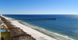 A beach with a long pier extending into the ocean, a sandy shoreline, and a clear blue sky.