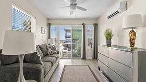 A modern living room with a grey sofa, a white floor lamp, and a grey cabinet. There is a ceiling fan, an air conditioning unit, and a view of a balcony with a green railing.