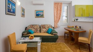 A cozy living room with a sofa, a coffee table, and a dining table with chairs. There are framed pictures on the wall, a window with curtains, and a bright yellow cabinet.