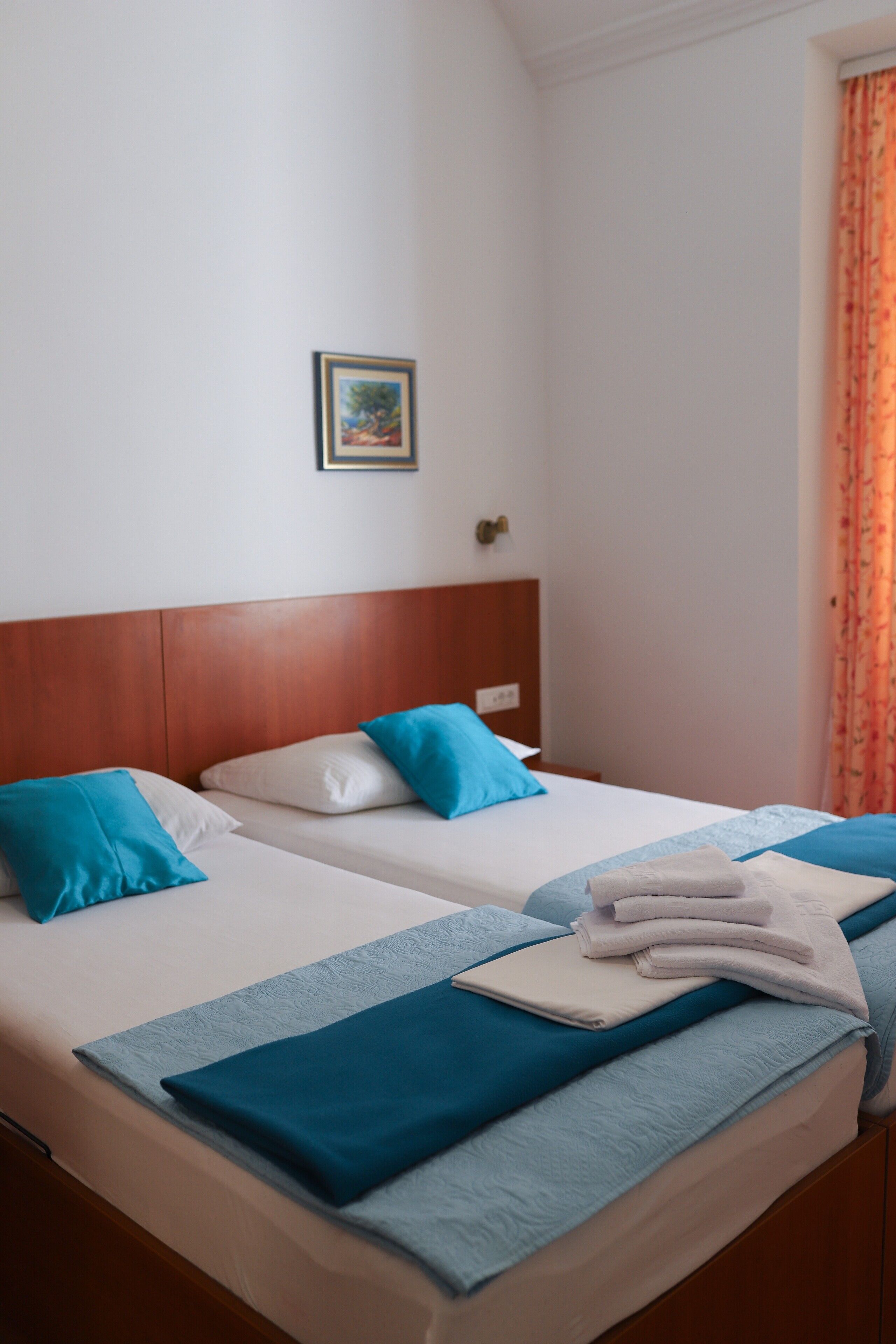 A hotel room with two beds, each with blue and white bedding, a wooden headboard, a framed picture on the wall, and a window with orange curtains.