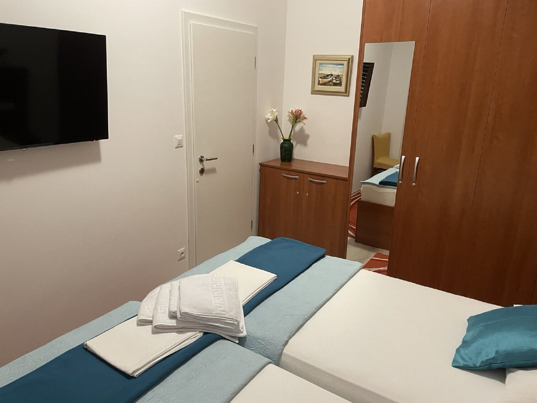 Un dormitorio con cama, televisor montado en la pared, armario y un pequeño aparador con un jarrón de flores.