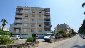 Un immeuble résidentiel à plusieurs étages, doté de balcons, d’un mur en pierre et bordé de voitures garées le long de la rue.