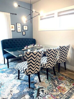 Uma sala de jantar com mesa de vidro, quatro cadeiras com padrões em zigue-zague, um sofá azul e um tapete estampado.