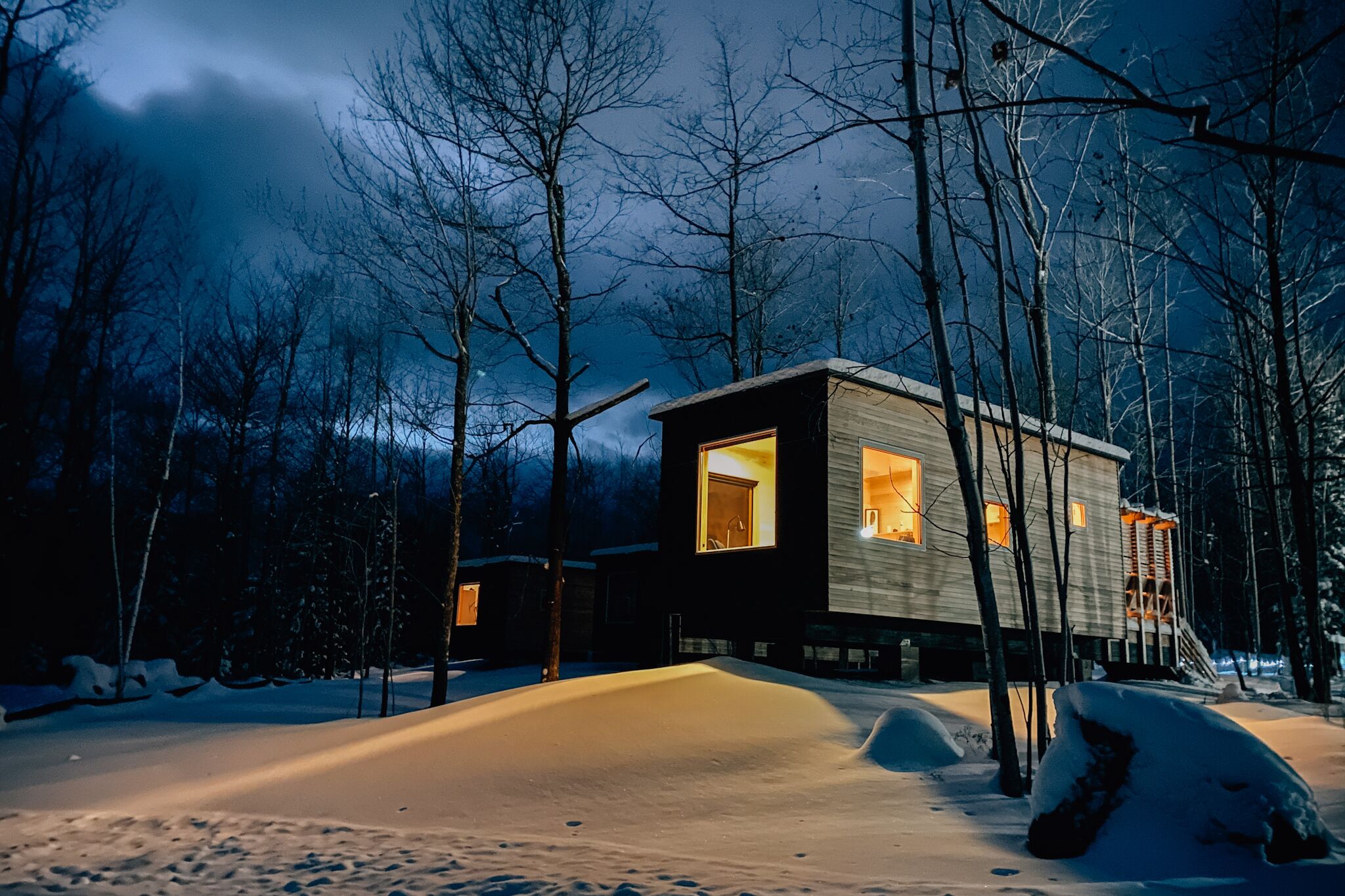 A modern cabin with large windows, surrounded by snow-covered ground and trees.