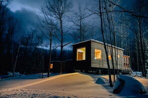 A modern cabin with large windows, surrounded by snow-covered ground and trees.