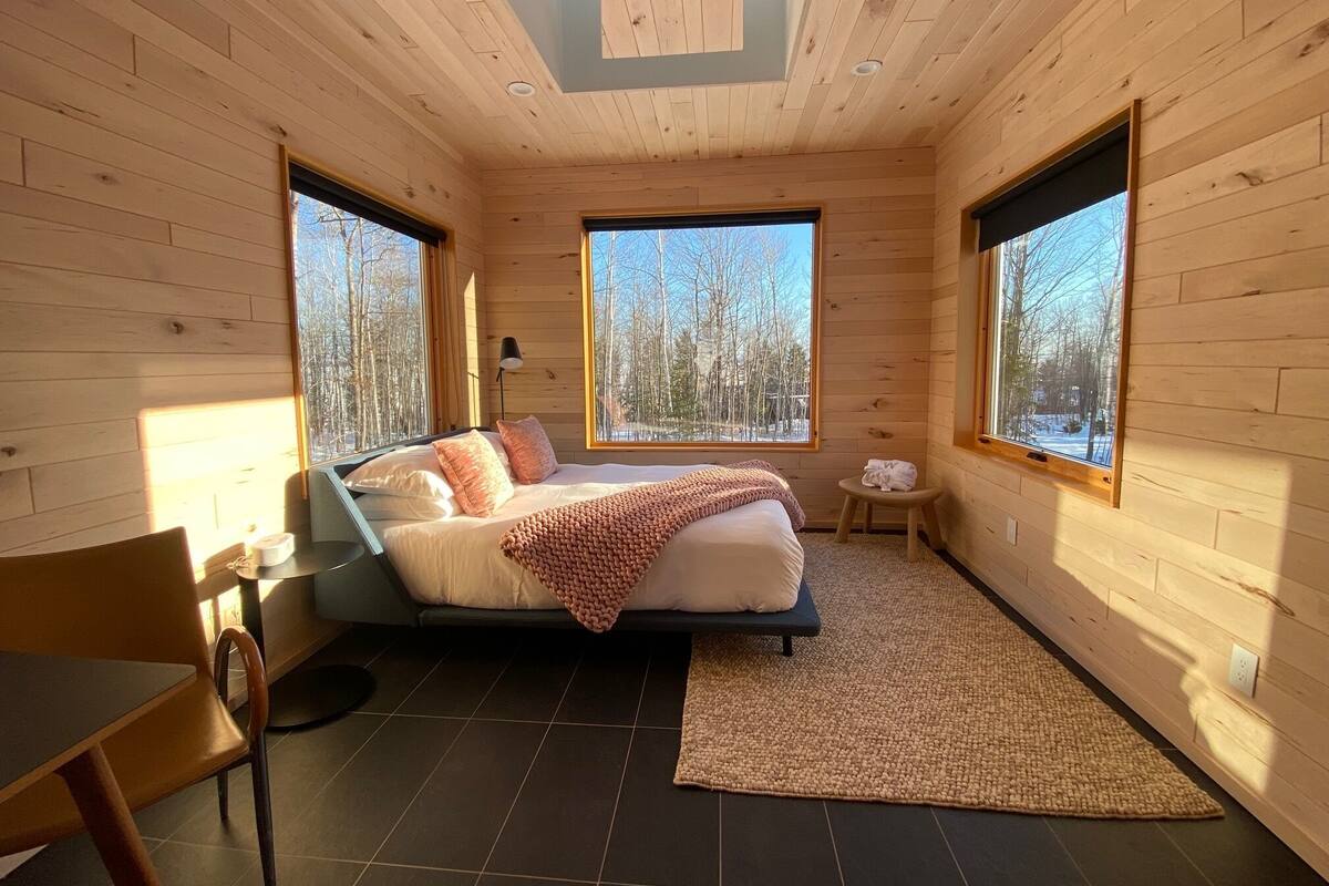 A cozy bedroom with a bed, a small table, and a chair. There are large windows offering a view of trees.