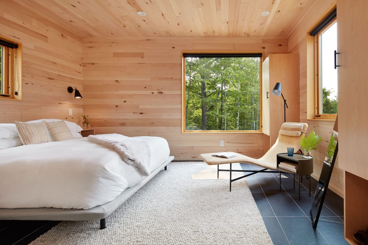 A modern bedroom with a large bed, a wooden ceiling, a window with a view of trees, and a chair with a side table.