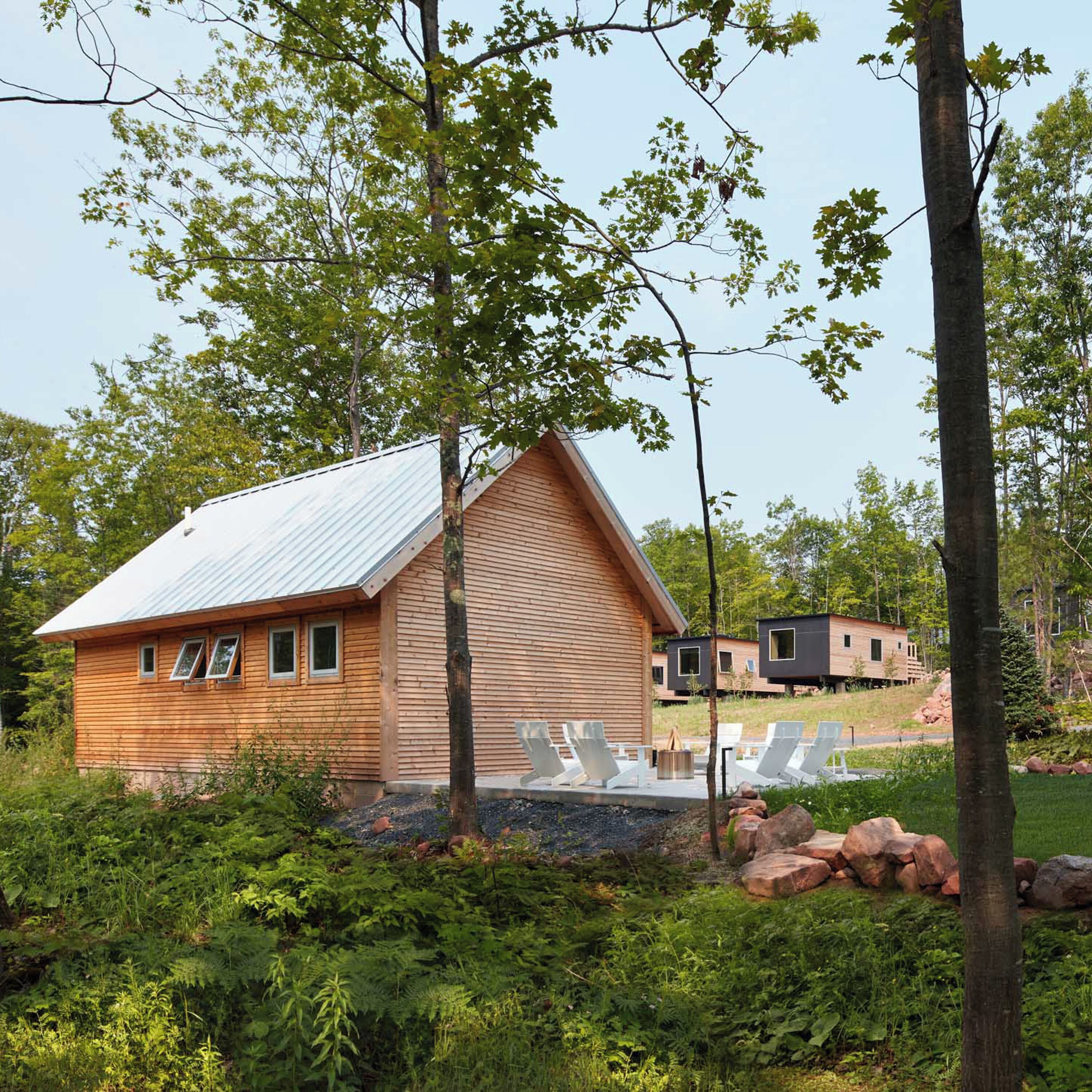 Une cabane en bois dotée d’un toit en pente, entourée d’une végétation luxuriante et comprenant un petit espace extérieur aménagé avec des chaises et un foyer.