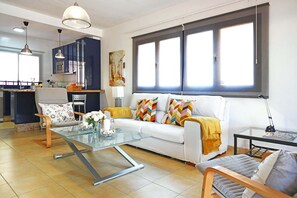 Ein modernes Wohnzimmer mit einem Glastisch, einem weißen Sofa mit bunten Kissen, einem Holzsesseln und einem Essbereich mit einer Küche im Hintergrund.