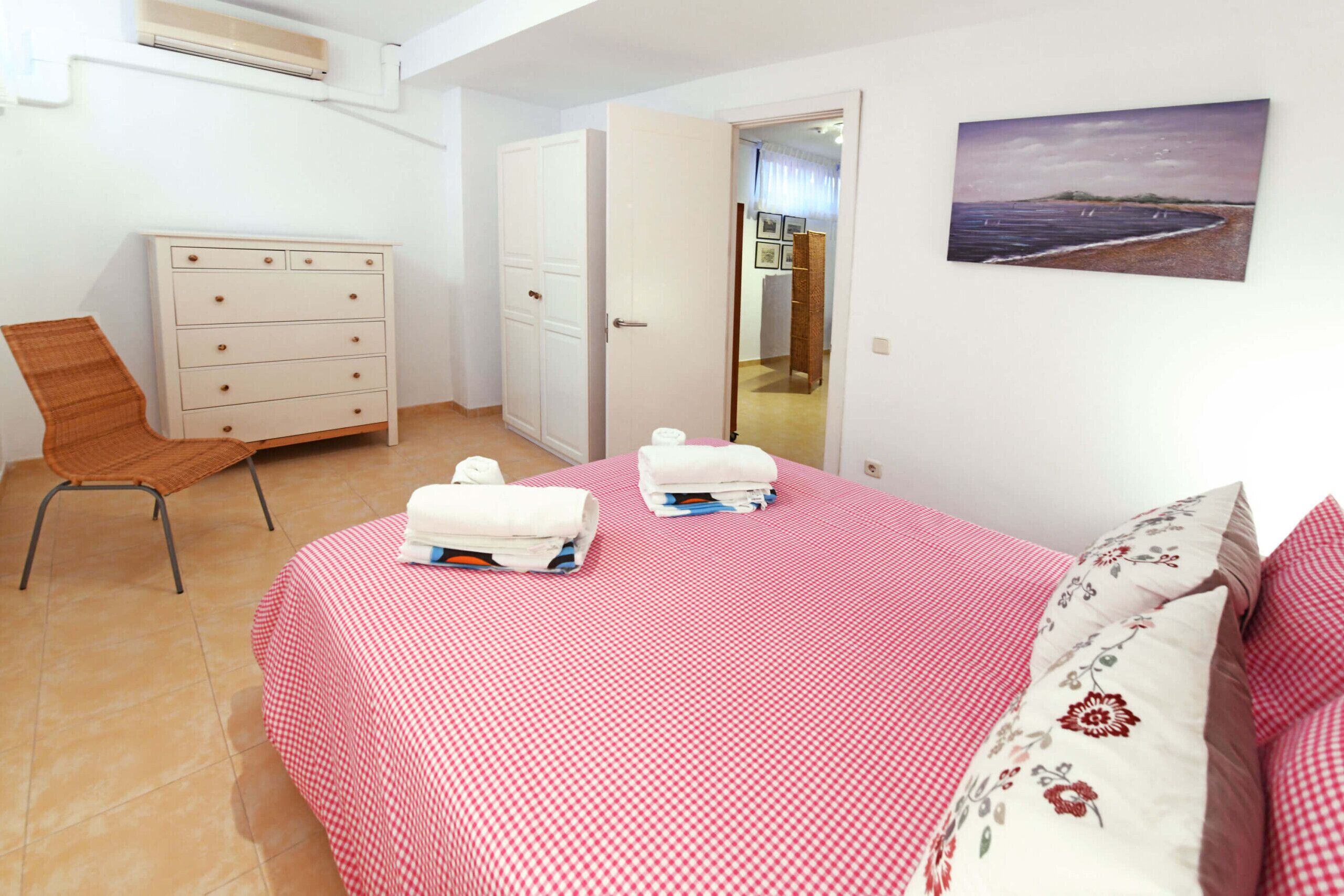 Une chambre avec une couvre-lit à carreaux, une chaise en bois, une commode blanche et un tableau représentant une scène de plage.