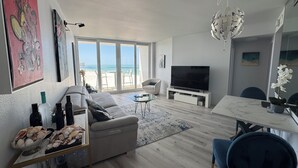 Ett modernt vardagsrum med en stor TV, bekväma sittplatser, en matplats och utsikt över stranden.