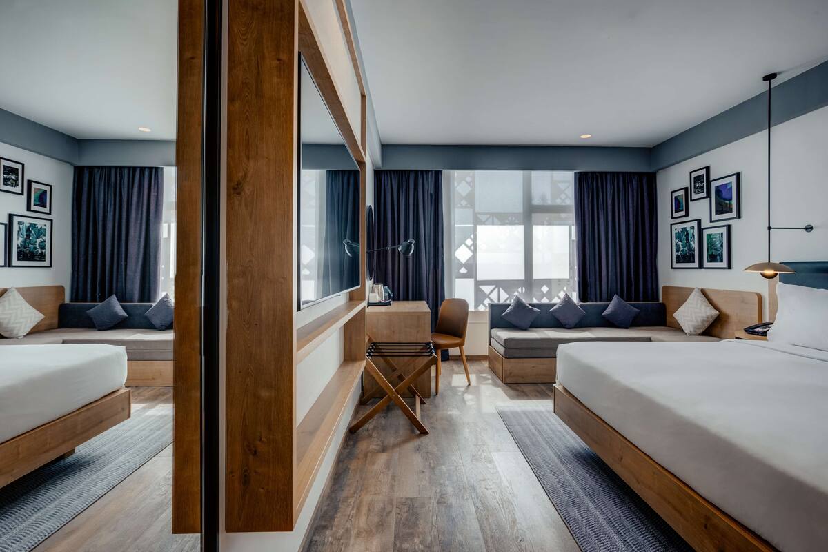 A modern hotel room with two beds, a sofa, a small table, and a chair. There are large windows with curtains, and artwork on the walls.
