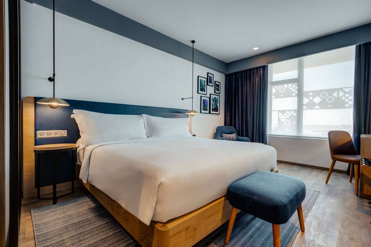 A modern hotel room with a large bed, bedside table, and a desk with a chair.