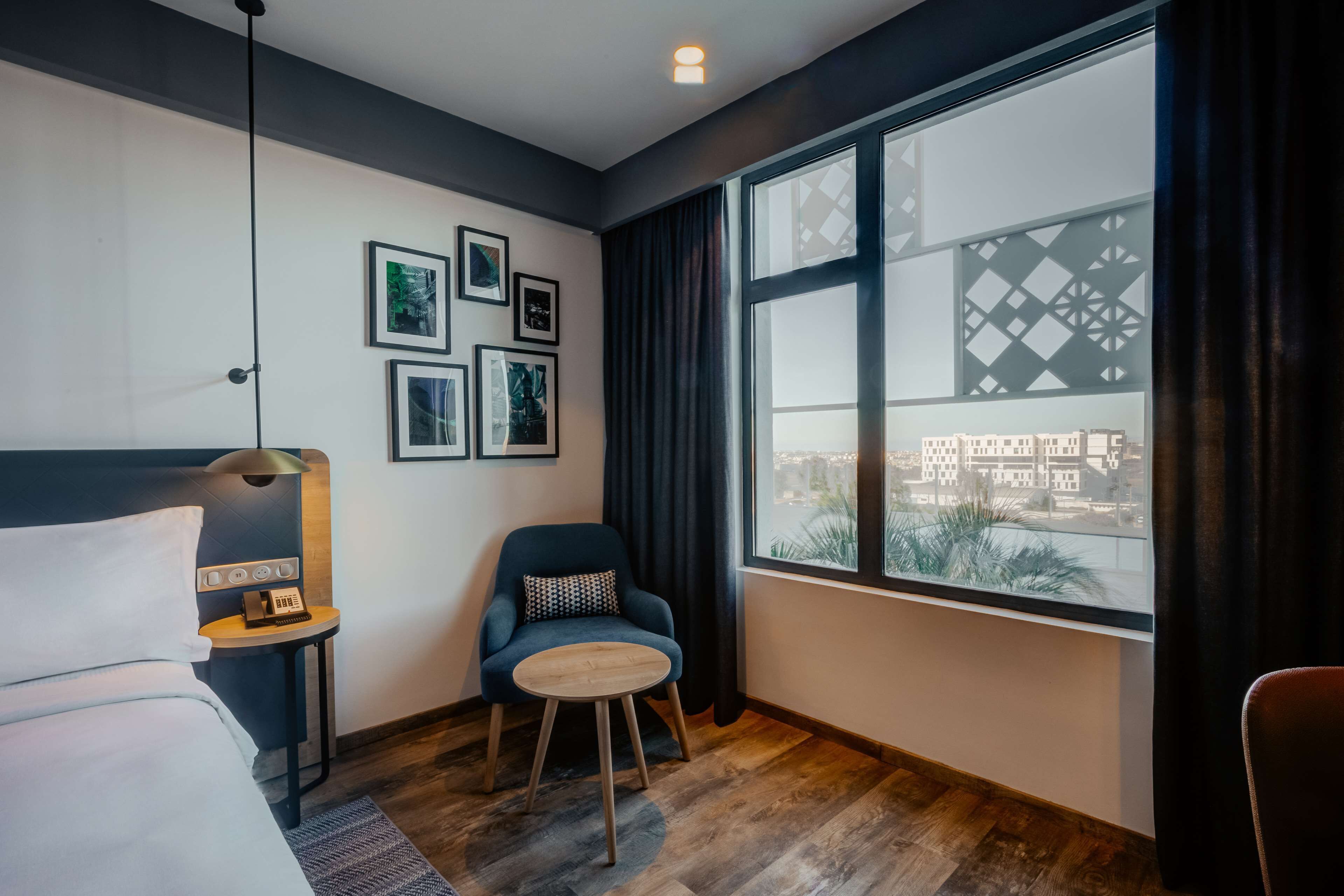 Ein modernes Hotelzimmer mit einem Bett, einem Schreibtisch mit Lampe, einem Sessel, einem runden Tisch und einem großen Fenster mit Blick auf die Stadt.