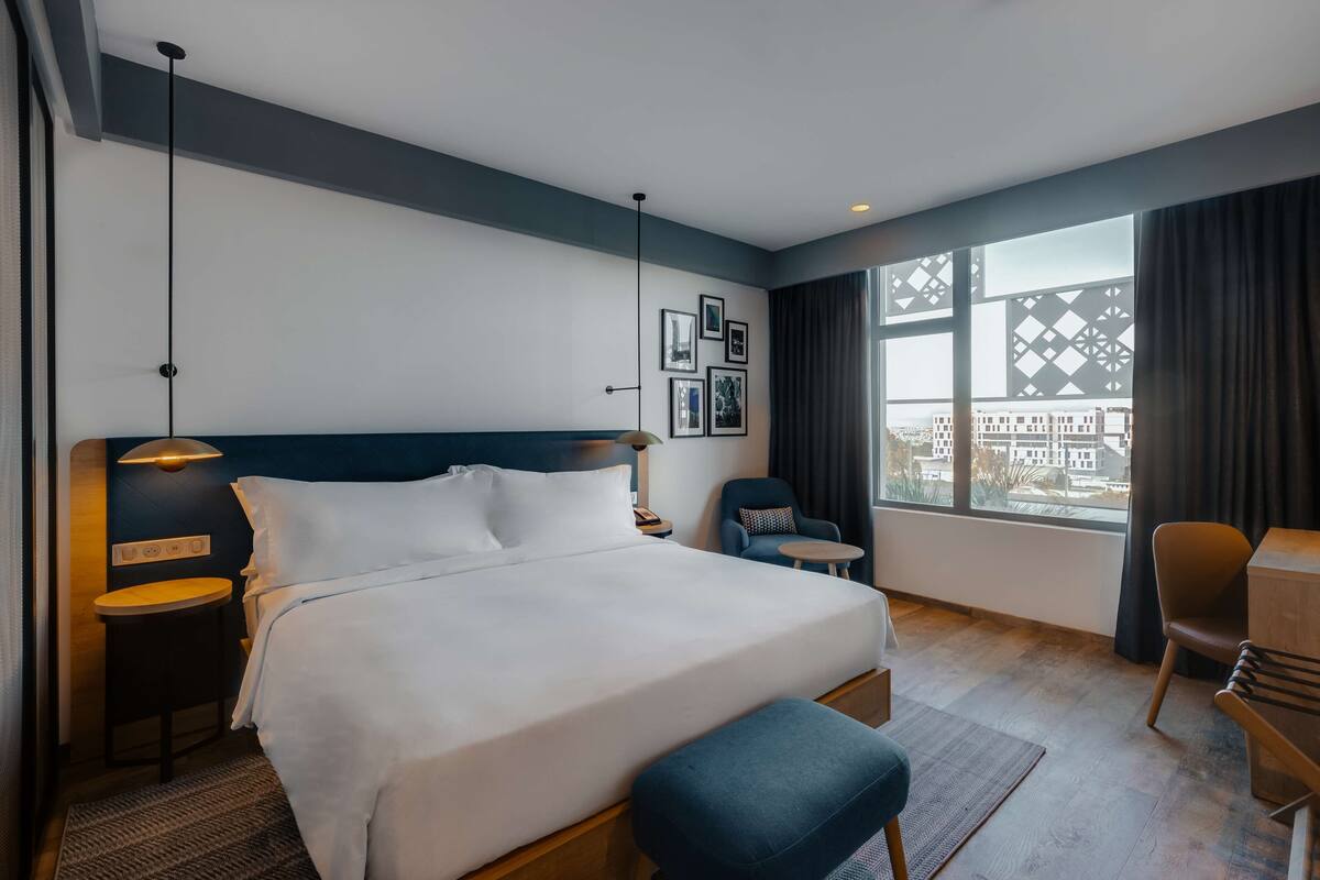 A hotel room with a large bed, a desk, a chair, and a view of buildings through the window.