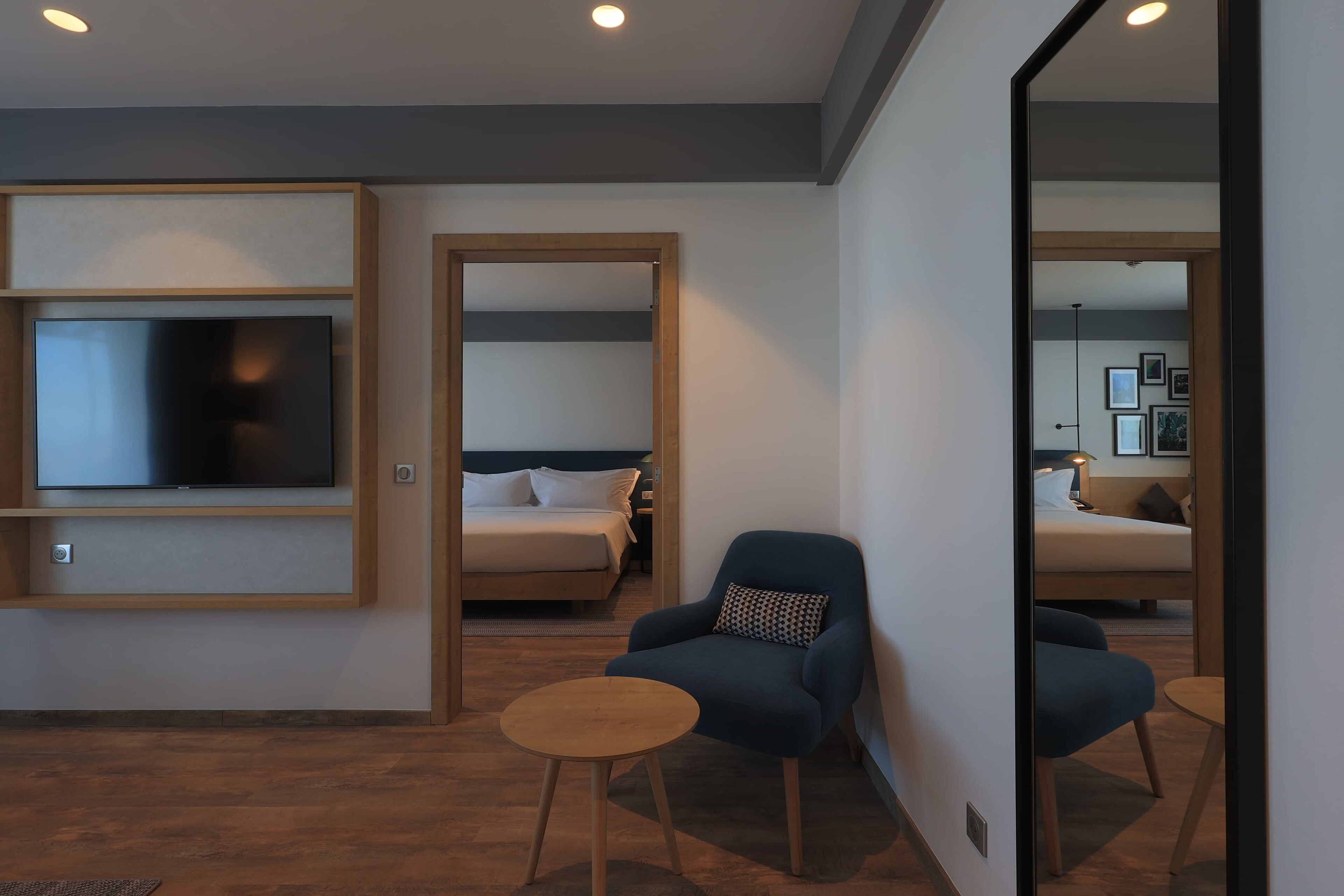A modern hotel room with a flat-screen TV, a bed, a blue armchair, and a small wooden table.