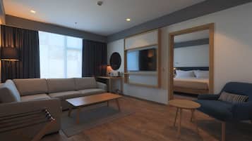 Habitación de hotel moderna con sofá, sillón, mesa de centro, televisor y cama con cabecera.