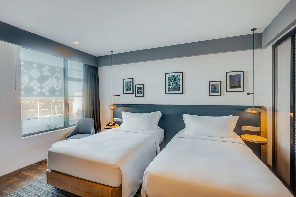 A hotel room with two beds, a large window with a view, and several framed pictures on the wall.