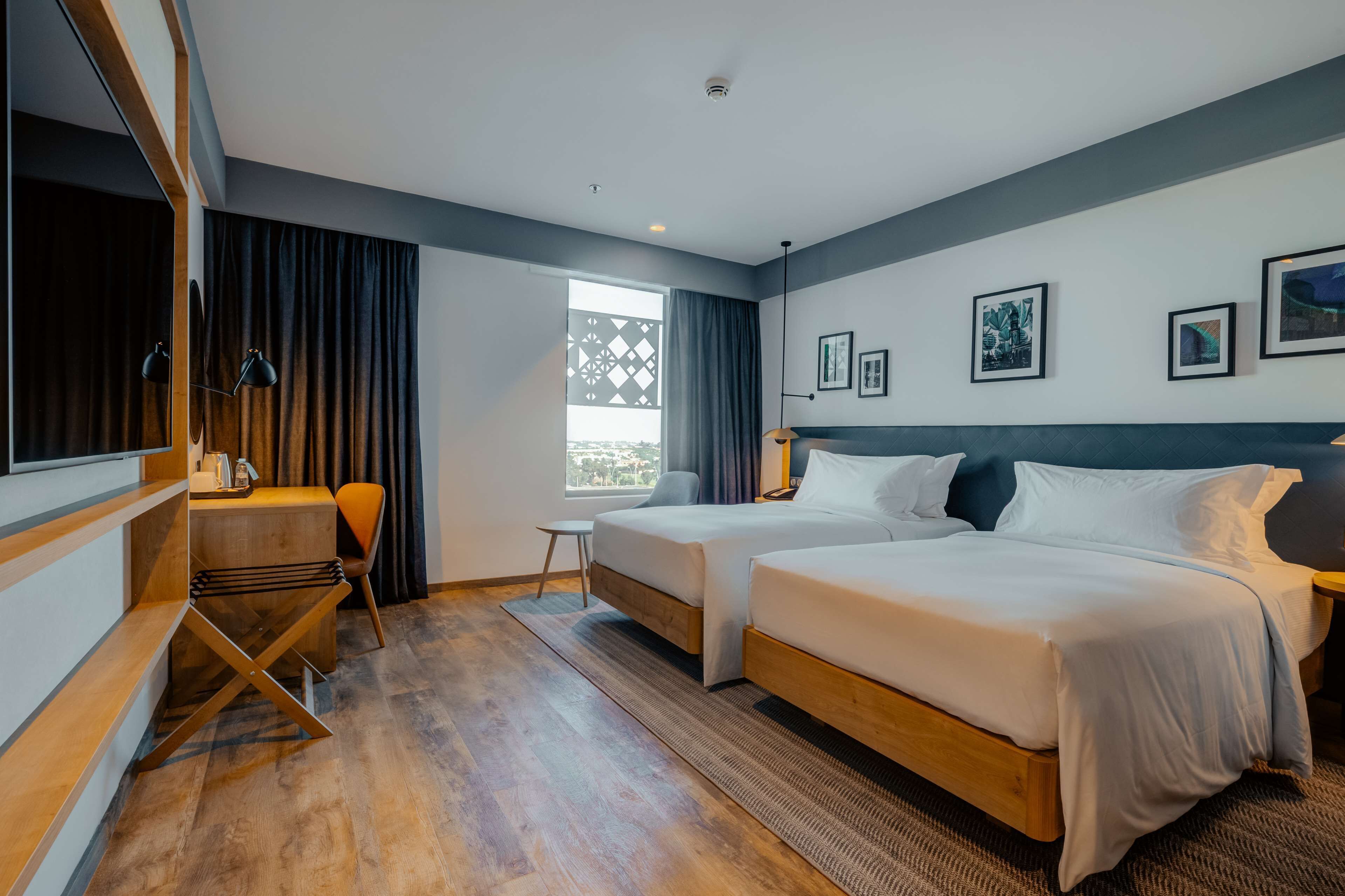 Quarto de hotel com duas camas, uma escrivaninha, uma cadeira, uma janela com cortinas e obras de arte nas paredes.