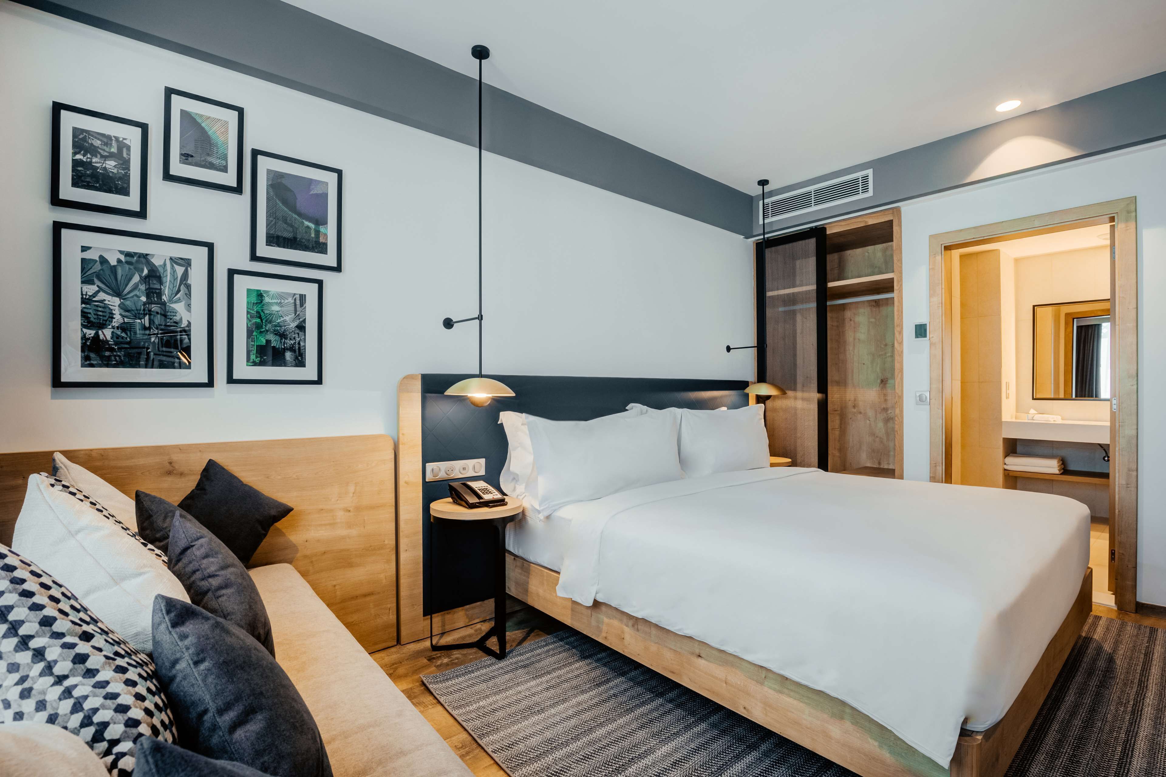 Una habitación de hotel moderna con una cama grande, un cabecero de madera, una mesita de noche con una lámpara, una pequeña mesa redonda y un televisor montado en la pared.