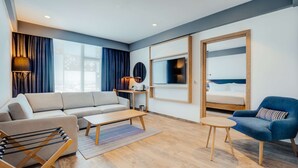 A modern living room with a sofa, coffee table, and armchair, a TV mounted on the wall, and a bed with a wooden frame. - Hilton Garden Inn Casablanca Sud (Casablanca)