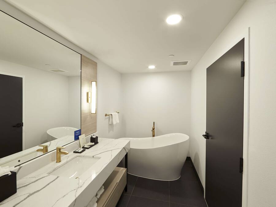 A modern bathroom with a large mirror, two sinks, a freestanding bathtub, and a dark door.