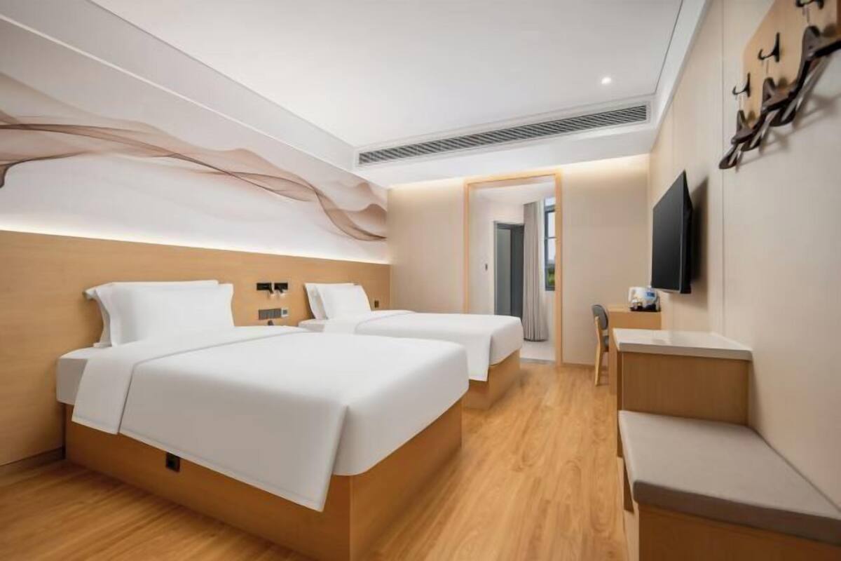 A modern hotel room with two beds, a flat-screen TV, a wooden bench, and a wall-mounted coat rack.