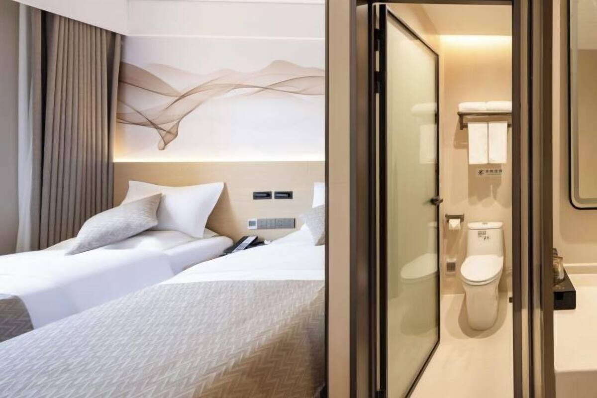 A hotel room with a bed, a bedside table, a wall-mounted light, and a bathroom with a toilet and sink.