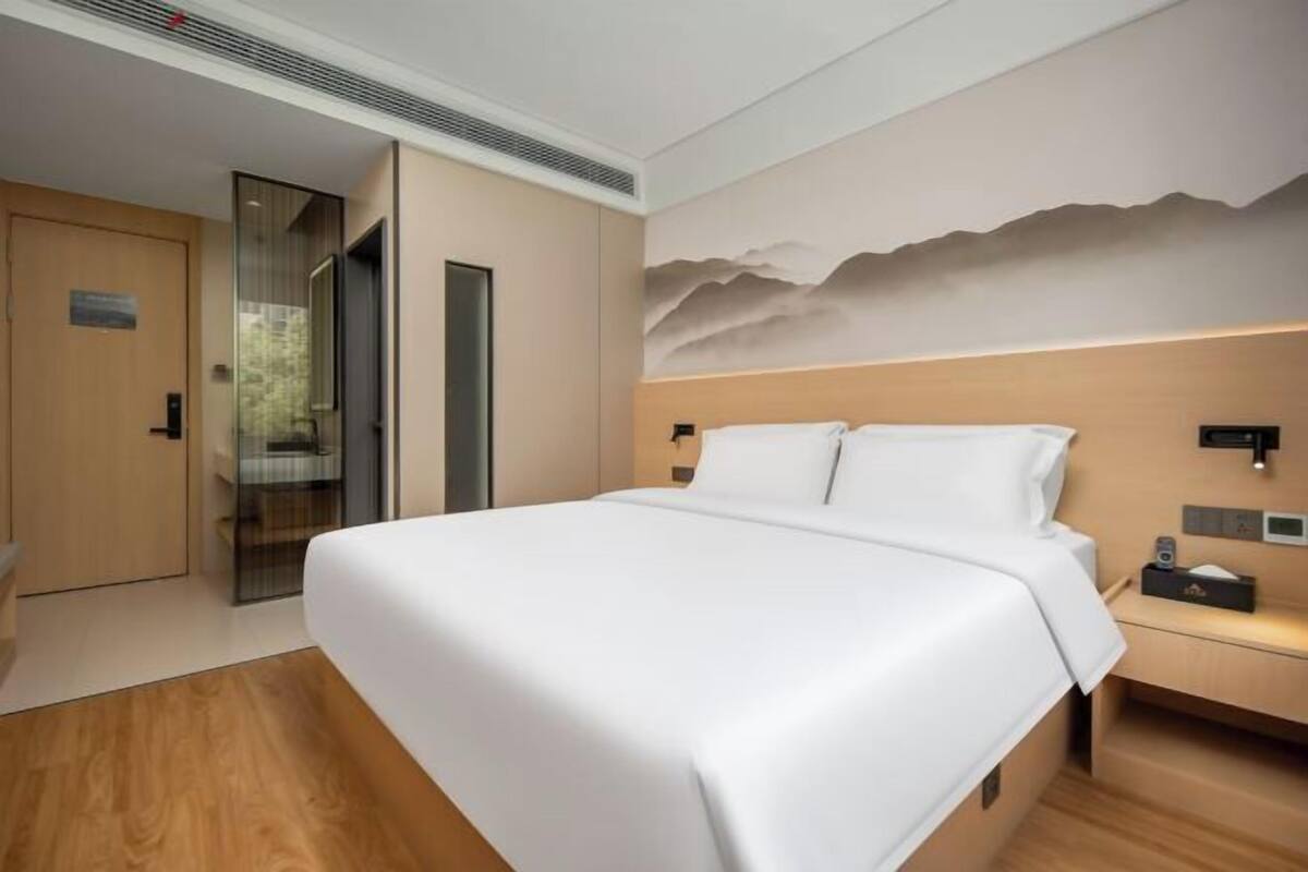 A modern hotel room with a large bed, a wooden floor, a wall-mounted nightstand, and a door leading to a bathroom.
