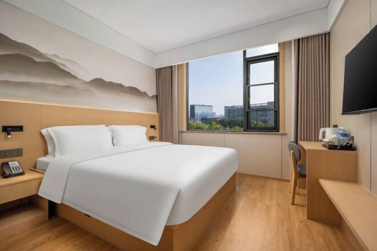 A hotel room with a bed, a desk, a chair, a TV, and a window with a city view.