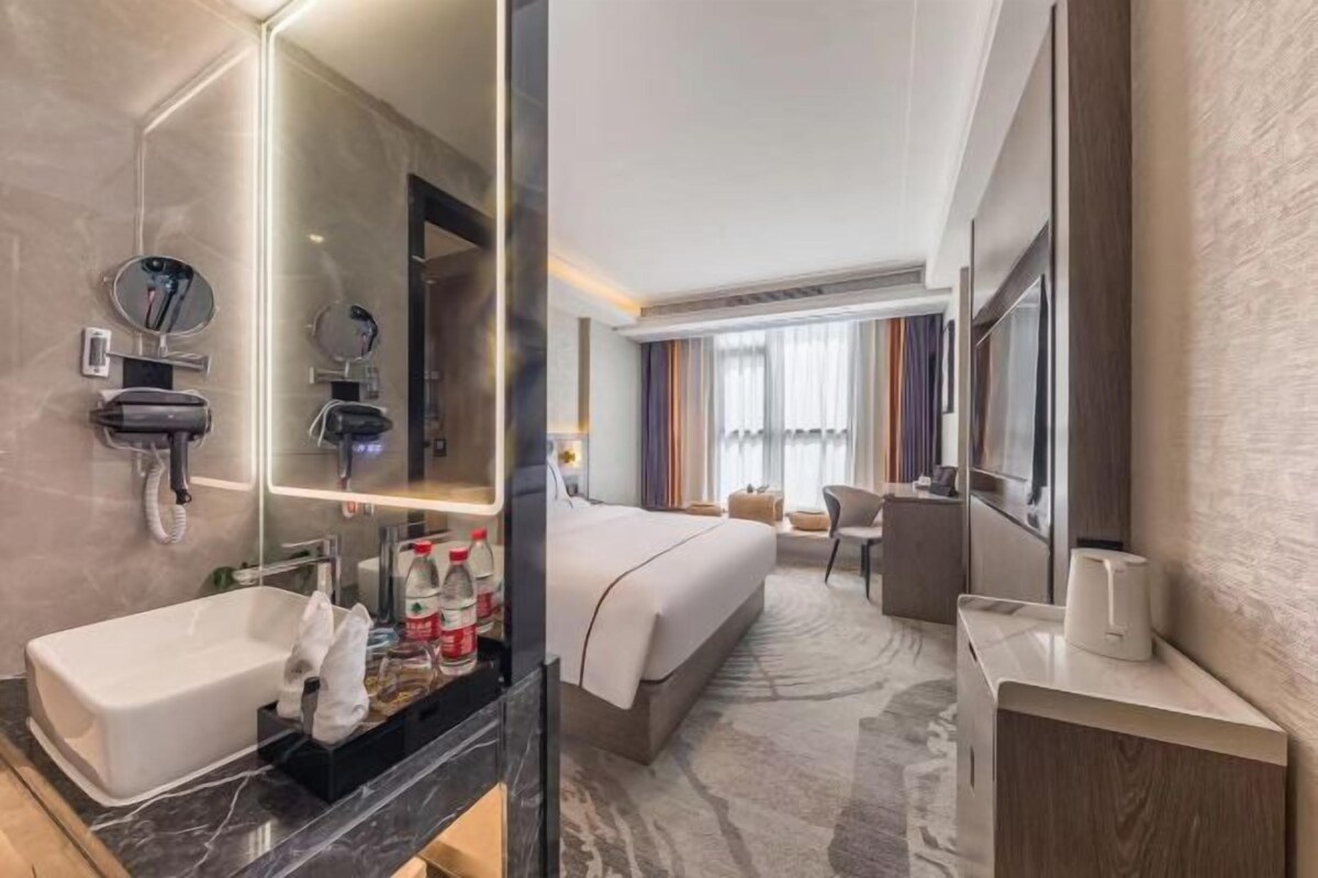 A modern hotel room with a large bed, a separate bathroom with a sink and mirror, a flat-screen TV, and a view of the city through large windows.