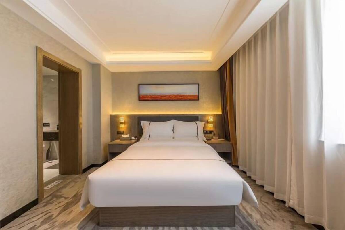 A hotel room with a large bed, bedside lamps, a painting on the wall, and a view of the outdoors through curtains.