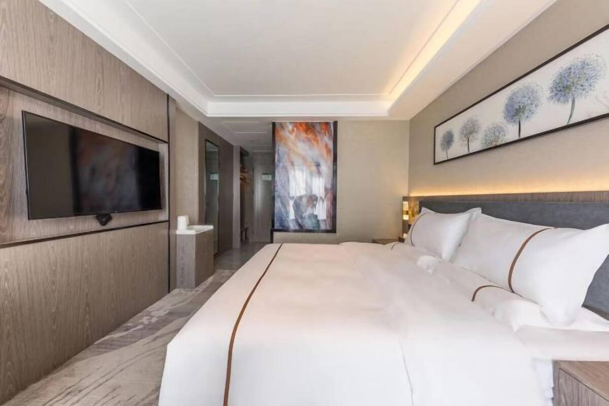 A modern hotel room with a large bed, a flat-screen TV, a painting on the wall, and a view of the hallway.