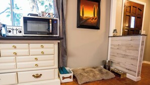 A kitchen area with a microwave, a white cabinet with gold handles, a framed picture on the wall, and a pet bed with a bone-shaped container. - The Paso House | Family Fun For All (Redding)
