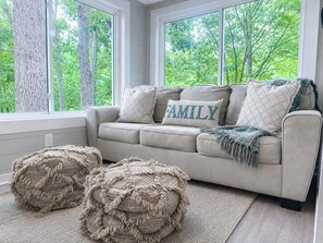 A beige sofa with a 'FAMILY' pillow, two round poufs, and a view of trees through large windows. - Sweeping Lake Views • Fire Pit • Hot Tub • Pets OK (Mooresville)