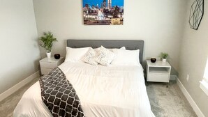 Une chambre à coucher bien rangée, avec un tableau représentant un paysage urbain, un plaid à motifs et deux tables de chevet ornées de plantes.