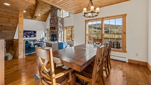 Uma ampla área de jantar com mesa e cadeiras de madeira, lareira de pedra e grandes janelas com vista para o exterior.