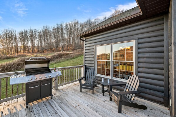 Una terraza de madera con una parrilla, dos sillas Adirondack y una mesa.