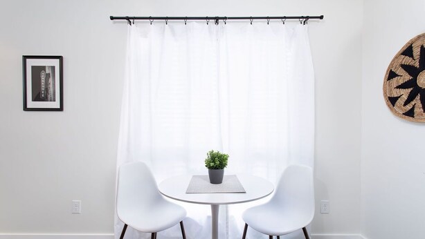 A room with a white curtain, a small table with a potted plant, two white chairs, and a round wall decoration. - City Condo with Outdoor Pool Near Broadway (Nashville)