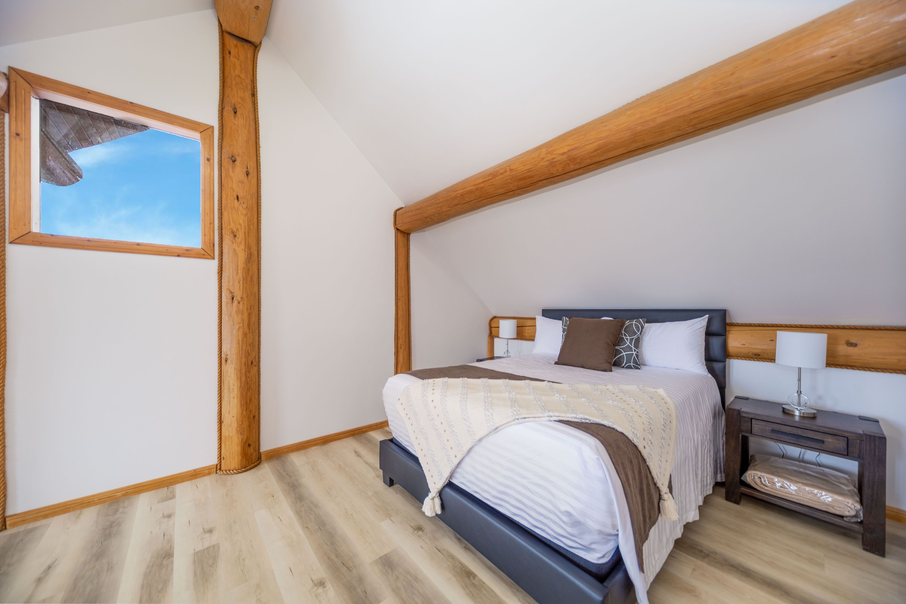 Un dormitorio con techo de madera, una cama con colcha blanca y marrón, una mesita de noche con lámpara y una ventana con vista al cielo.