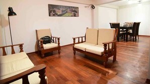 Ein Zimmer mit Holzboden, zwei Sofas aus Holz, einem Esstisch mit Stühlen, einer Stehlampe und einem gerahmten Bild an der Wand.