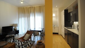 Sala de estar moderna com TV, sofá e área de jantar. Cozinha com armários escuros e bancada branca visível.