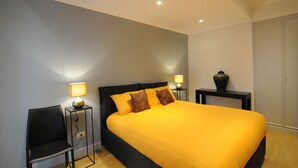 Quarto com cama grande, duas mesas de cabeceira, uma cadeira e um criado-mudo com abajur.