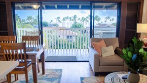 A room with a wooden dining table, chairs, a sofa with cushions, a potted plant, and a view of palm trees through a balcony door. - Kiahuna Plantation Ocean View with AC, REMODELED, 1 Bedroom, Sleeps 4 (Poipu)